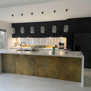 May include: A modern kitchen island with a white countertop and a gold metal backsplash. The island has a built-in sink and a set of pendant lights hanging above it.