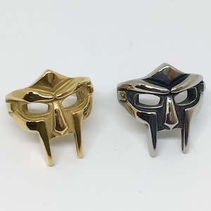 Puede incluir: Dos anillos de metal con un diseño de máscara. Un anillo es dorado y el otro es plateado.
