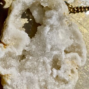 May include: A white crystal geode pendant with a gold chain. The geode has a large opening that reveals a cluster of smaller crystals inside.