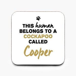 May include: White square coaster with black text that reads "This human belongs to a Cockapoo called Cooper". The text "Cooper" is in gold. A black paw print is above the text.