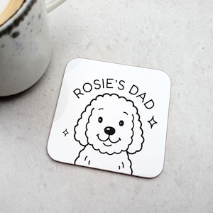 May include: White square coaster with a black and white illustration of a poodle and the text "Rosie's Dad". The coaster has rounded corners and two small starbursts on either side of the dog's head.