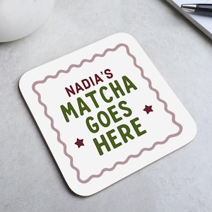 May include: Square white coaster with a wavy border and the text "NADIA'S MATCHA GOES HERE" in green and burgundy. Two burgundy star shapes are on the bottom. The coaster is on a light grey surface.