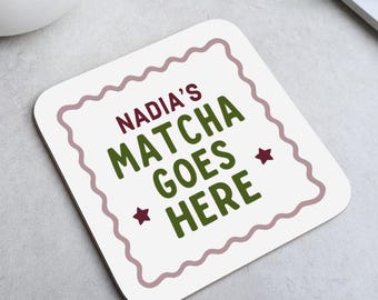 May include: Square white coaster with a wavy border and the text "NADIA'S MATCHA GOES HERE" in green and burgundy. Two burgundy star shapes are on the bottom. The coaster is on a light grey surface.