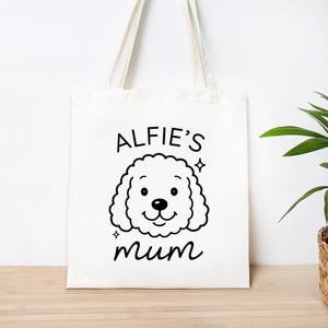 May include: A white tote bag with a black illustration of a dog's face and the text "ALFIE'S mum". The dog illustration has a curly coat and a smiling expression. The bag has long, cream-colored handles.