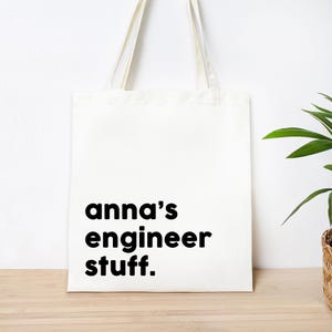 May include: A white canvas tote bag with two long shoulder straps. The front of the bag features the black printed text "anna's engineer stuff." in a simple, bold, sans-serif font. The bag has a rectangular shape and a clean, flat design.