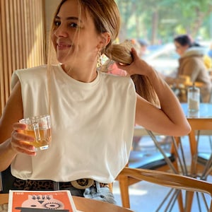 May include: A woman wearing a white sleeveless shirt with a leopard print belt sits at a table with a book titled "OTOMOTIK PORTAKAL" by Anthony Burgess. She is holding a glass of tea.