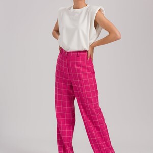 May include: A woman is wearing a white sleeveless top with padded shoulders and pink plaid wide-leg pants.