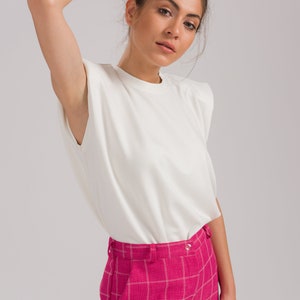 May include: A woman wearing a white, short-sleeved t-shirt with padded shoulders and pink plaid pants.