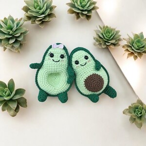 Puede incluir: Dos peluches de aguacate tejidos a crochet con caras sonrientes. El aguacate de la izquierda tiene un lazo morado. Ambos están sobre un fondo blanco con suculentas verdes.