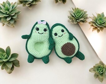 Könnte beinhalten: Zwei gehäkelte Avocado-Plüschtiere mit lächelnden Gesichtern. Die Avocado links hat eine lila Schleife. Beide befinden sich auf einem weißen Hintergrund mit grünen Sukkulenten.