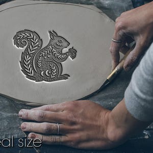 Op de afbeelding: Een lichtgrijs kleistuk met een gedetailleerd eekhoornontwerp dat een eikel vasthoudt. De eekhoorn is omgeven door decoratieve bladerpatronen. De tekst "Not real size" en "Size depends on your choice" staat onderaan de afbeelding.