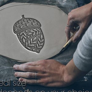May include: A gray clay acorn stamp with a detailed leaf and star design. The text "Not real size, Size depends on your choice" is at the bottom. The stamp is on a gray surface.