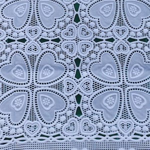 Può includere: Tessuto di pizzo bianco con un motivo a cuori ripetuto. Il tessuto ha un design delicato e intricato.