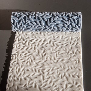 Op de afbeelding: Een witte deegroller met een reliëfpatroon van bladeren op het oppervlak. De deegroller is gemaakt van plastic en heeft een blauwe handgreep.