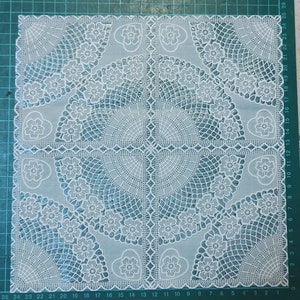 May include: White lace doily with a delicate floral and heart design. The doily has a circular pattern in the center and a square shape.