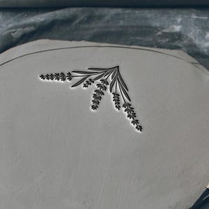 Peut inclure: Une pièce d'argile grise avec un motif de lavande est en cours de fabrication. Le texte "Not real size, Size depends on your choice" est visible. L'artisan utilise un outil pour façonner l'argile, soulignant le caractère artisanal du produit.