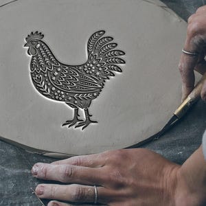 Puede incluir: Una pieza de arcilla gris claro con un diseño detallado de gallo. El texto "Not real size" y "Size depends on your choice" es visible. El gallo se está tallando con una herramienta, mostrando la naturaleza artesanal del producto.