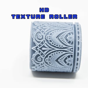 Pode incluir: Um rolo de textura cinza com um padrão intrincado e repetitivo. O rolo cilíndrico apresenta um design detalhado de formas florais e geométricas. O texto "HD TEXTURE ROLLER" é exibido em azul acima do rolo.