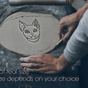Pode incluir: Uma peça plana de argila cinzenta de formato oval com um design em relevo de uma cabeça de gato com olhos em forma de abóbora e uma teia de aranha numa orelha. O texto "Not real size" e "Size depends on your choice" está impresso em branco.