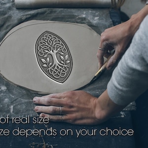Op de afbeelding: Een lichtgrijs kleistuk met een Keltisch levensboomontwerp wordt gemaakt. De ovale klei heeft de woorden "Not real size" en "Size depends on your choice" in witte tekst.