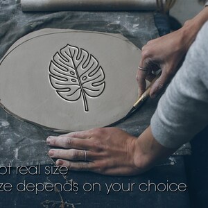 May include: A flat, oval-shaped piece of gray clay featuring a deeply impressed monstera leaf design in the center. The surface shows clear, clean lines from the stamp, and the text "Not real size. Size depends on your choice" is visible.