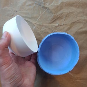 Può includere: Un oggetto bianco, liscio e circolare a forma di ciotola tenuto in aria accanto a un oggetto azzurro, strutturato e circolare a forma di ciotola con un bordo ruvido e irregolare. Entrambi gli oggetti sono aperti e cavi, mostrando le superfici interne.