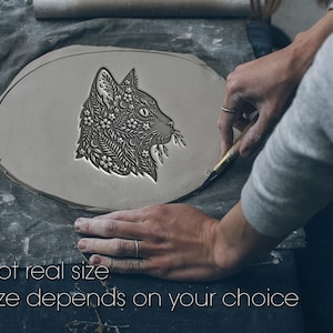 Può includere: Un pezzo di argilla grigia, piatto e ovale, con un'intricata incisione in rilievo di un profilo di testa di gatto. Il design del gatto è riempito con dettagli floreali, foglie e lune crescenti. Testo: Not real size. Size depends on your choice.