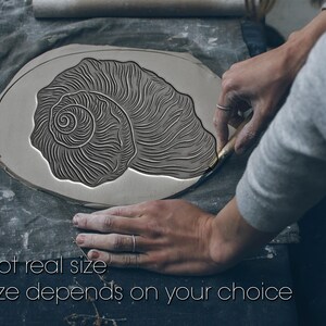 May include: A gray, oval-shaped clay piece with a detailed seashell design being carved. The text "Not real size" and "Size depends on your choice" is visible. The image shows the process of creating a ceramic art piece.
