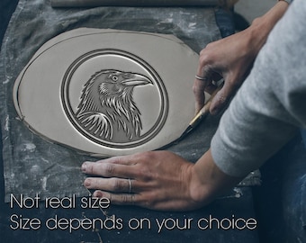 May include: A gray, oval-shaped ceramic piece with a detailed raven design in a circular frame. The text "Not real size" and "Size depends on your choice" is visible. The piece is being worked on by a person.