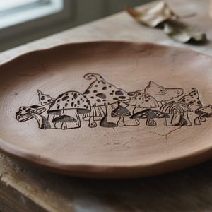 以下が含まれることがあります： テラコッタ色の丸い陶器の皿で、様々なキノコのハンドドローイングイラストが描かれています。キノコは様々な形と模様で一列に並んでいます。皿の縁は少し不均一です。