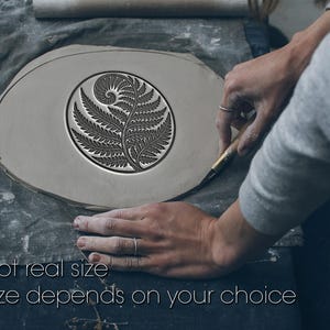 Op de afbeelding: Een lichtgrijs kleistuk met een varenontwerp in het midden. De tekst "Not real size" en "Size depends on your choice" staat onderaan. Een persoon gebruikt een gereedschap om de klei te vormen.