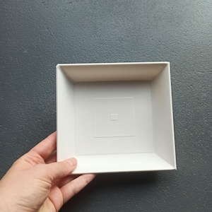 May include: A square, off-white plastic tray with slanted sides. The interior base features a raised square border design with a small, centered square indentation. The tray has a smooth, matte finish and a clean, geometric structure.