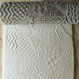 May include: A textured clay roller with various geometric and organic patterns is shown above a sheet of clay with the same patterns. The roller is gray and cylindrical, and the patterns include zigzags, triangles, and swirls. The clay sheet is white.