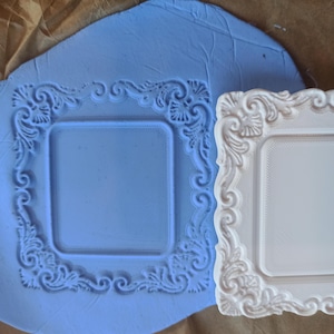 Könnte beinhalten: Eine flexible hellblaue Silikonform mit quadratischer Mitte und verziertem Schnörkelrand, platziert neben einem weißen Abguss aus derselben Form, der das identische, komplizierte und erhabene dekorative Rahmendesign zeigt.