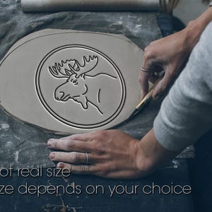 May include: A light gray, oval-shaped clay piece with a circular moose design is being crafted. The moose is depicted in a minimalist, black line drawing. The text "Not real size" and "Size depends on your choice" is visible.