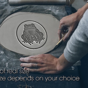 May include: A light gray ceramic piece with a detailed frog design inside a circle. The frog is decorated with stars and crescent moons. The text "Not real size" and "Size depends on your choice" is at the bottom of the image.