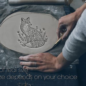 Op de afbeelding: Een lichtgrijs, ovaal kleistuk met een gedetailleerd vossendesign en bloemaccenten. De vos is ingewikkeld bewerkt. De tekst "Not real size" en "Size depends on your choice" staat onderaan. Een persoon bewerkt het stuk.