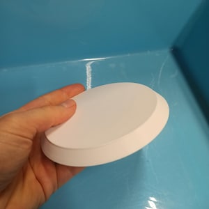 Pode incluir: Um objeto branco, redondo, impresso em 3D com uma borda ligeiramente elevada. O objeto é segurado em uma mão contra um fundo azul claro. Parece um prato pequeno ou uma tampa.