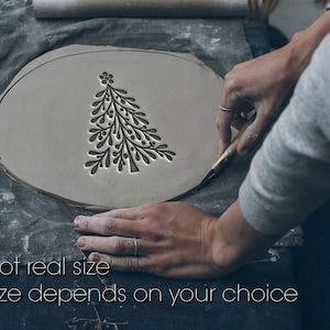 May include: A light gray clay piece with a carved Christmas tree design. The text "Not real size" and "Size depends on your choice" is visible. The clay is being worked on a gray cloth surface.