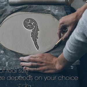 Puede incluir: Una pieza de arcilla gris claro con un diseño detallado y arremolinado que se está tallando. El texto "Not real size" y "Size depends on your choice" está en la parte inferior. La arcilla está sobre un paño gris.
