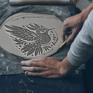 Könnte beinhalten: Ein detailliertes, graues Keramikstück mit einem Kolibri-Design wird geschnitzt. Die Flügel des Vogels sind ausgebreitet, und das Design weist komplizierte Muster auf. Die Hände des Künstlers sind sichtbar und halten ein Werkzeug zum Schnitzen des Tons. Das Stück liegt auf einem grauen Tuch.