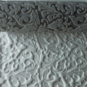 Può includere: Primo piano di un mattarello grigio con un motivo floreale decorativo. Il mattarello viene utilizzato per creare un motivo in rilievo su una superficie bianca.