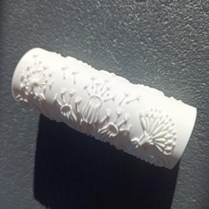 Op de afbeelding: Een witte cilindrische verfroller met een verhoogd, reliëfpatroon van paardenbloemzaadhoofdjes en stengels. De textuur is direct in het oppervlak van de roller gegoten, wat een herhalend botanisch ontwerp creëert voor decoratief schilderwerk.