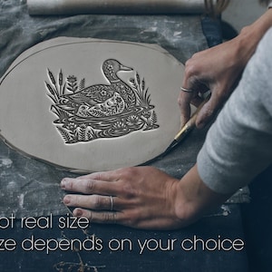 Op de afbeelding: Een plat, ovaalvormig stuk grijze klei met een gedetailleerde, in reliëf gemaakte illustratie van een eend die in het water zwemt, omringd door riet en bloemen. De tekst 'Not real size' en 'Size depends on your choice' staat onderaan.