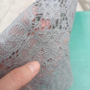 Può includere: Un pezzo di tessuto di pizzo grigio con un intricato motivo floreale viene sollevato, rivelando una superficie turchese sottostante. Il pizzo presenta disegni dettagliati di fiori, foglie e motivi a vortice, creando una trama delicata ed elegante.