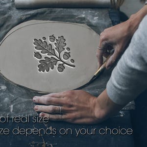Op de afbeelding: Een lichtgrijs kleistuk met een eikenblad- en eikelontwerp dat wordt gesneden. De tekst "Not real size" en "Size depends on your choice" is zichtbaar. De klei ligt op een grijs oppervlak.
