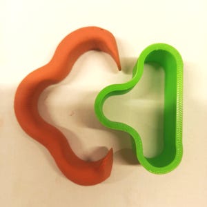 Può includere: Due formine per biscotti in plastica. Una è di forma curva arancione brillante, l'altra è un rettangolo verde arrotondato con un'indentazione curva. Le formine sono probabilmente per la cottura o l'artigianato.