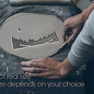 Peut inclure: Une pièce d'argile grise de forme ovale avec un paysage sculpté représentant des arbres, une maison, une lune et des étoiles. Le texte "Not real size" et "Size depends on your choice" est en bas. Une personne utilise un outil pour sculpter l'argile.