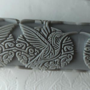 May include: Close-up of a gray rolling pin with a repeating hummingbird design. The hummingbird is depicted in a circular pattern with detailed wing and body features. The design is raised, creating a textured surface.