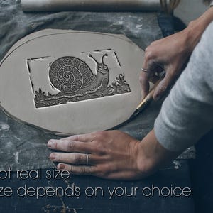 May include: A flat, circular piece of grey clay featuring a stamped rectangular design of a snail with a detailed, swirling shell pattern. The text "Not real size. Size depends on your choice" is printed in white at the bottom of the image.